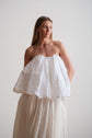 Model wearing a strapless white top with gathered volume and a flowing skirt, shown against a neutral backdrop.