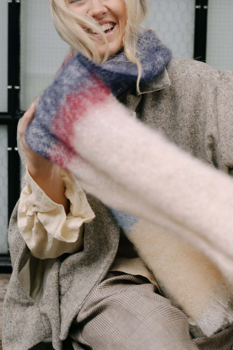 Woman smiling in a textured coat while holding a soft striped scarf close to the camera in natural daylight.