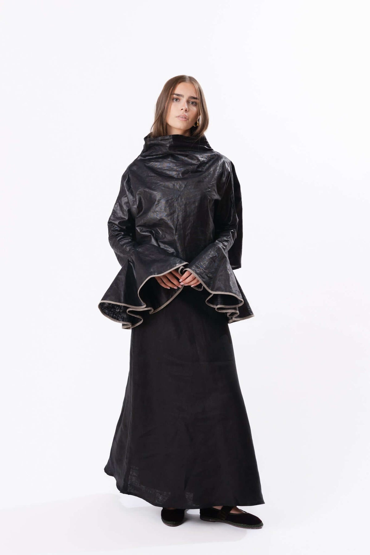 Model wearing a dark sculptural cape over a long skirt, creating a dramatic silhouette against a plain backdrop.