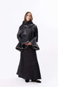Model wearing a dark sculptural cape over a long skirt, creating a dramatic silhouette against a plain backdrop.