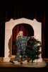 Model in a checked outfit standing on a theatre stage beside a small tree and painted arch set in warm stage lighting.