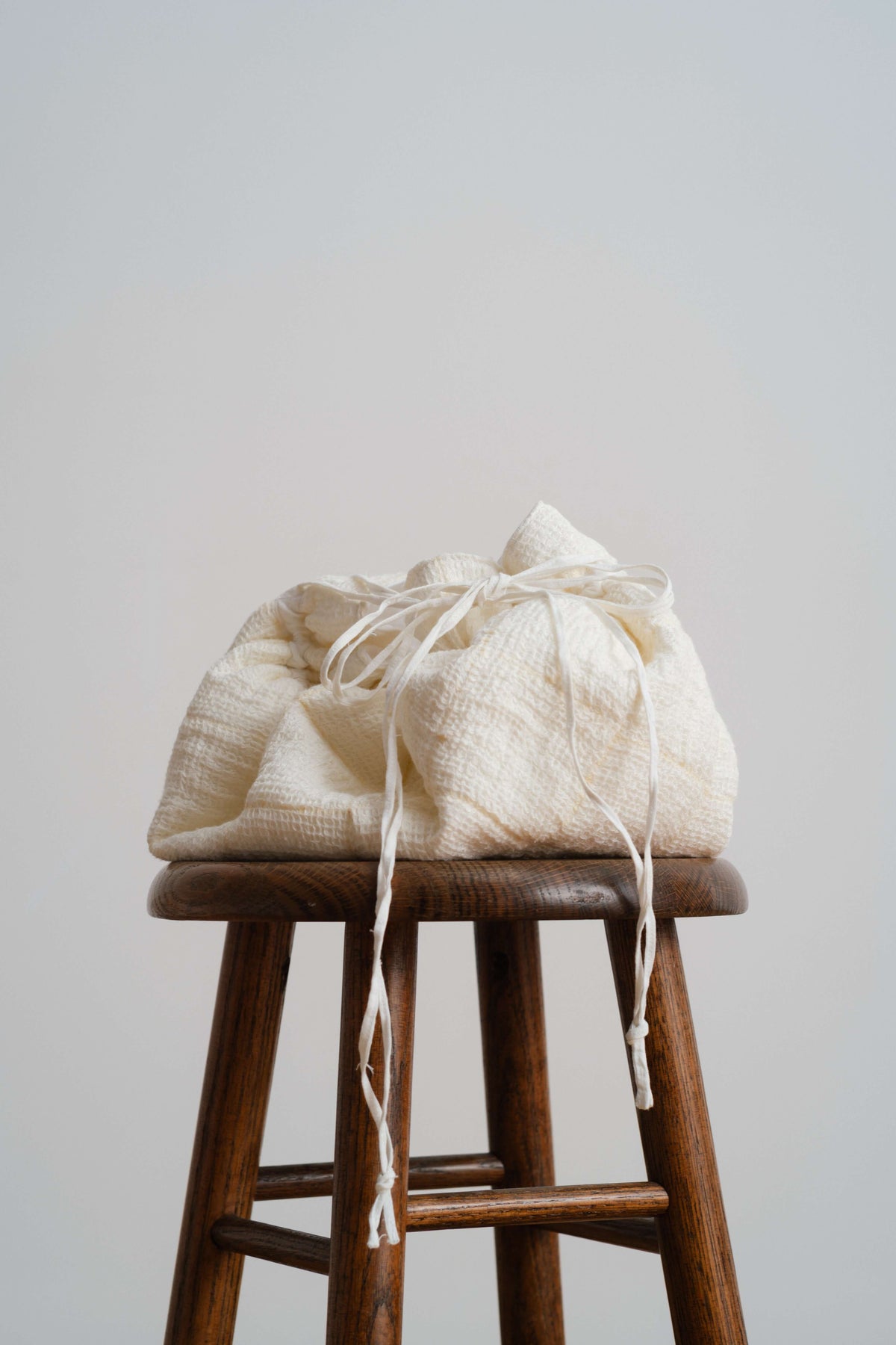 Stack of folded light garments tied with ribbon and placed on a wooden stool against a plain wall in soft daylight.