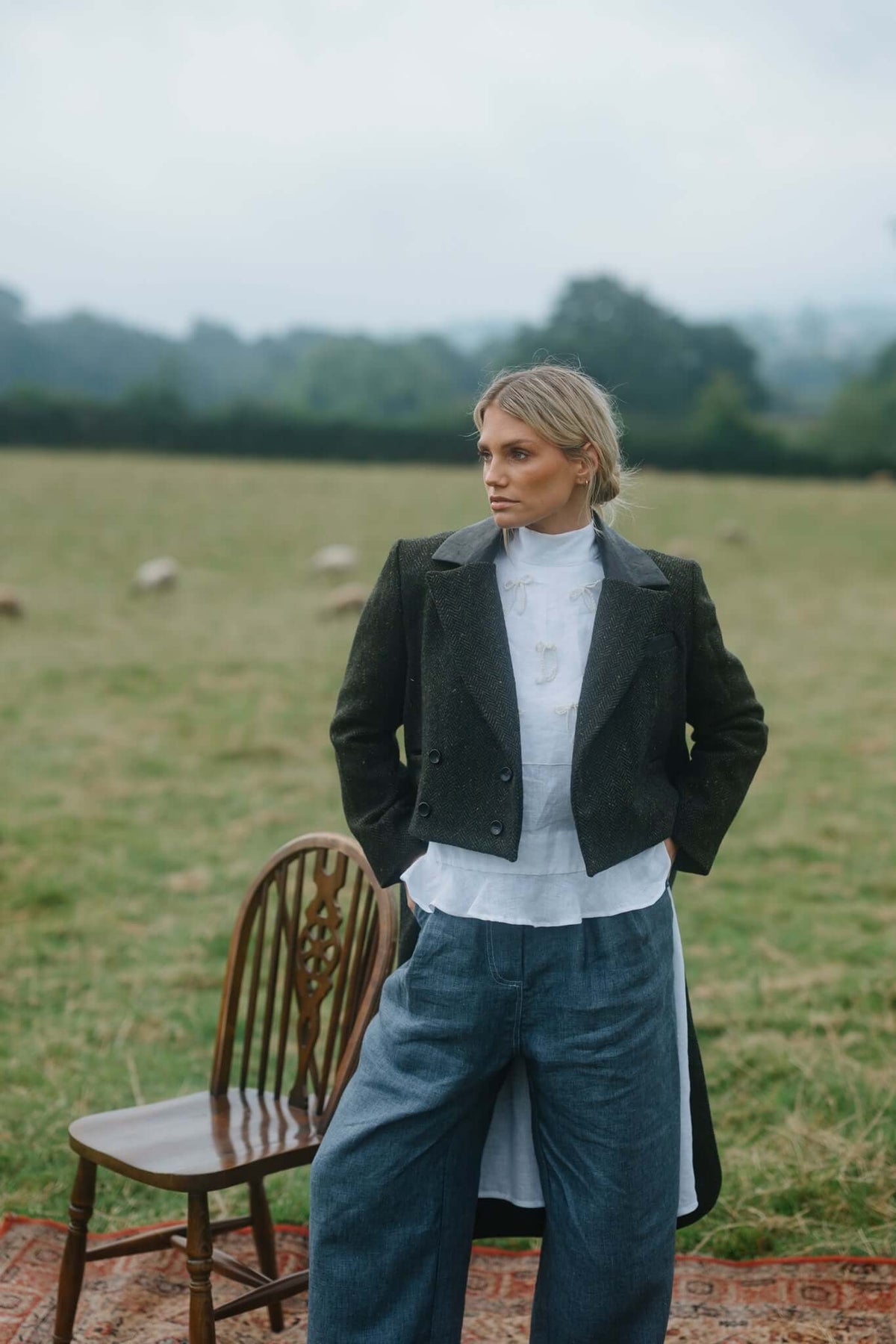 Woman in a dark tailored jacket and cropped trousers standing outdoors beside a chair in an open field.