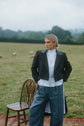 Woman in a dark tailored jacket and cropped trousers standing outdoors beside a chair in an open field.