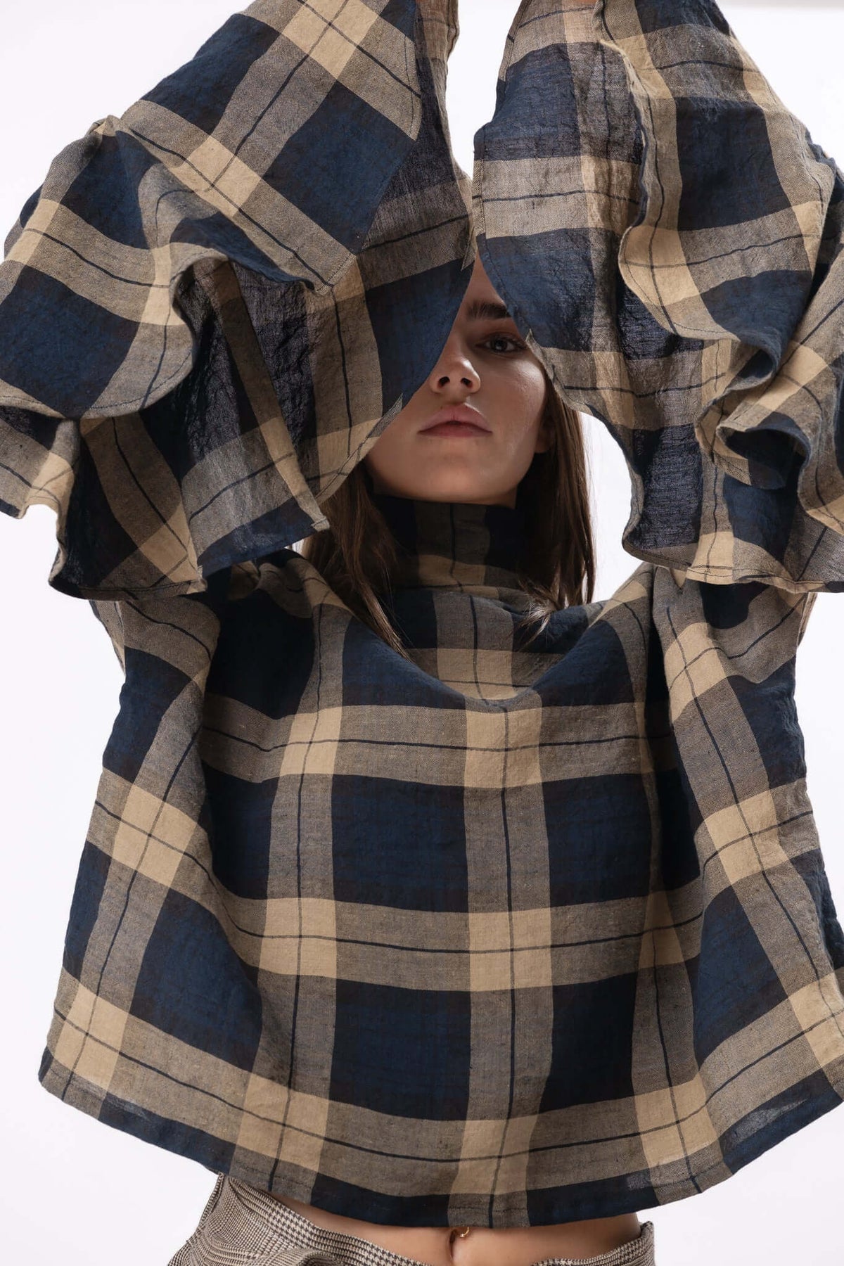 Close view of a checked dress with oversized sleeves raised overhead, highlighting structure, scale and movement.