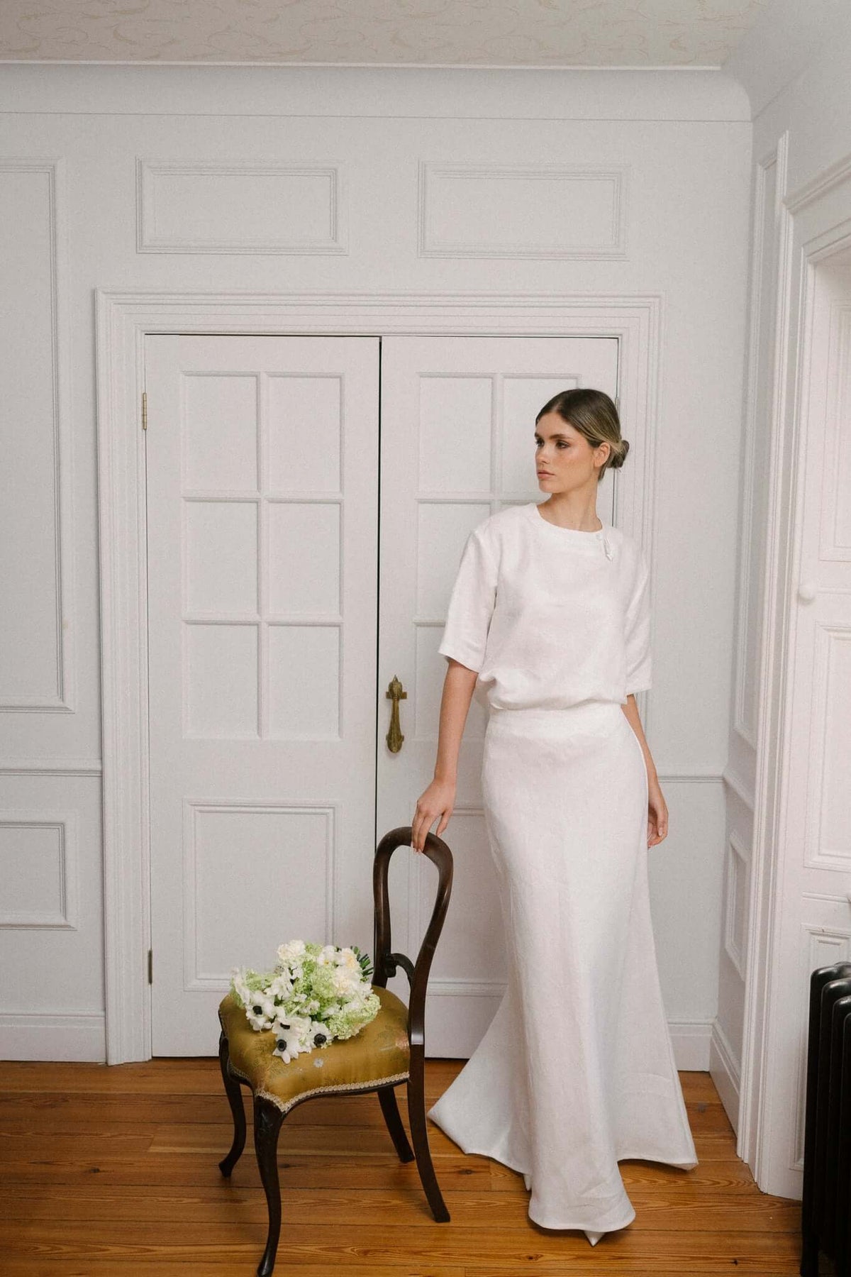 Woman in a white blouse and long skirt standing beside a chair indoors, photographed in soft window light.