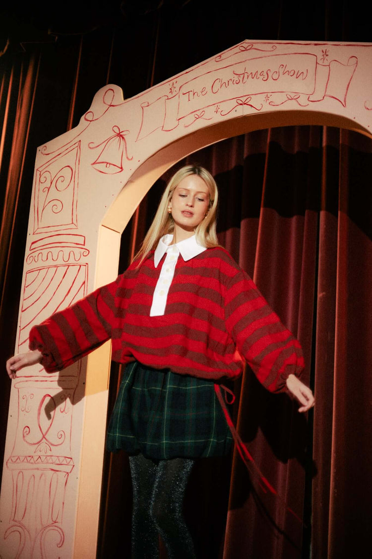 Model in a red striped knit and dark skirt standing on a theatre stage beneath a painted arch and red curtain.
