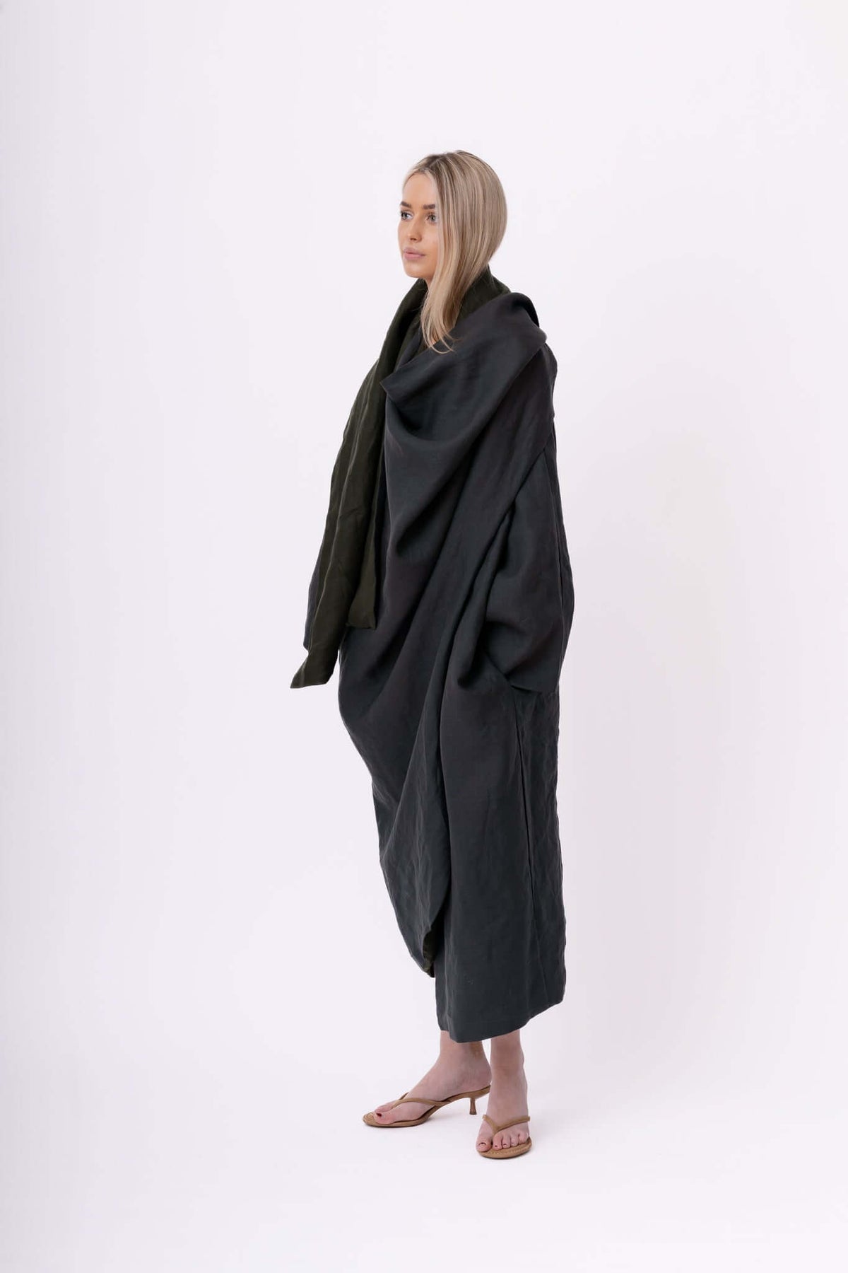 Model wrapped in a dark draped shawl or coat, standing barefoot against a pale studio background in a studio setting.