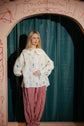 Woman in a printed blouse and checked trousers standing before a curtain and painted theatre arch in soft stage light.