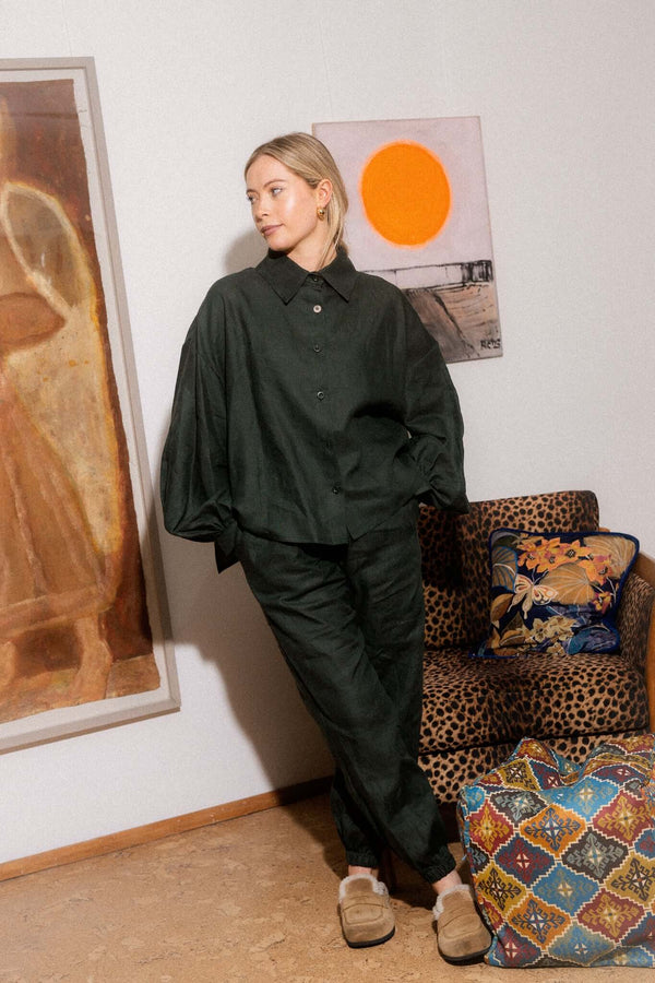 Woman in a dark relaxed outfit leaning beside artwork and patterned furniture in an interior setting.