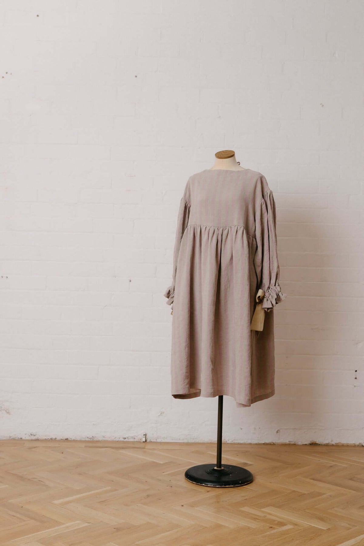 Neutral long-sleeved dress displayed on a mannequin against a bare wall and wooden floor in a studio interior.