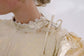Close detail of a cream high-neck top with lace trim, ribbon tie and softly textured metallic fabric.