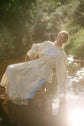 Woman seated outdoors in a flowing cream dress, photographed in hazy sunlight beside water and foliage.