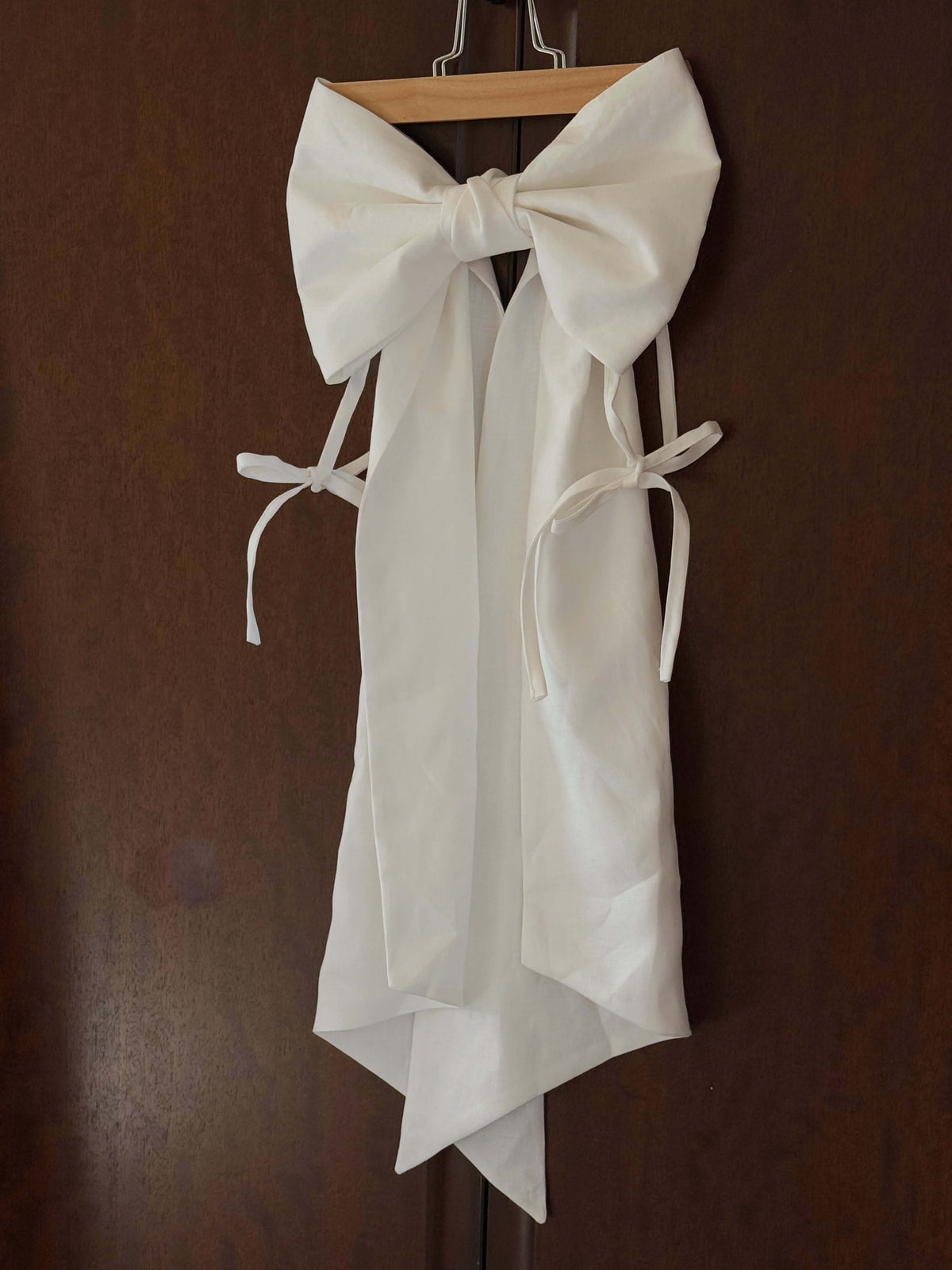 White dress or apron with oversized bow hanging on a wooden door, shown as a styled product detail on a dark wooden door.