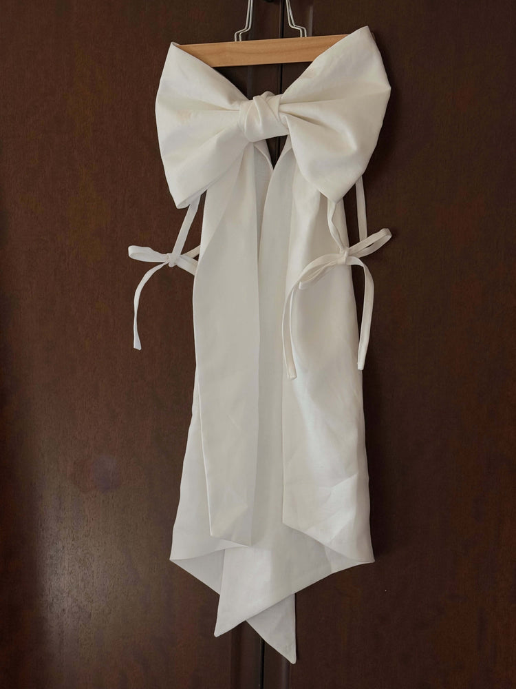 White dress or apron with oversized bow hanging on a wooden door, shown as a styled product detail on a dark wooden door.
