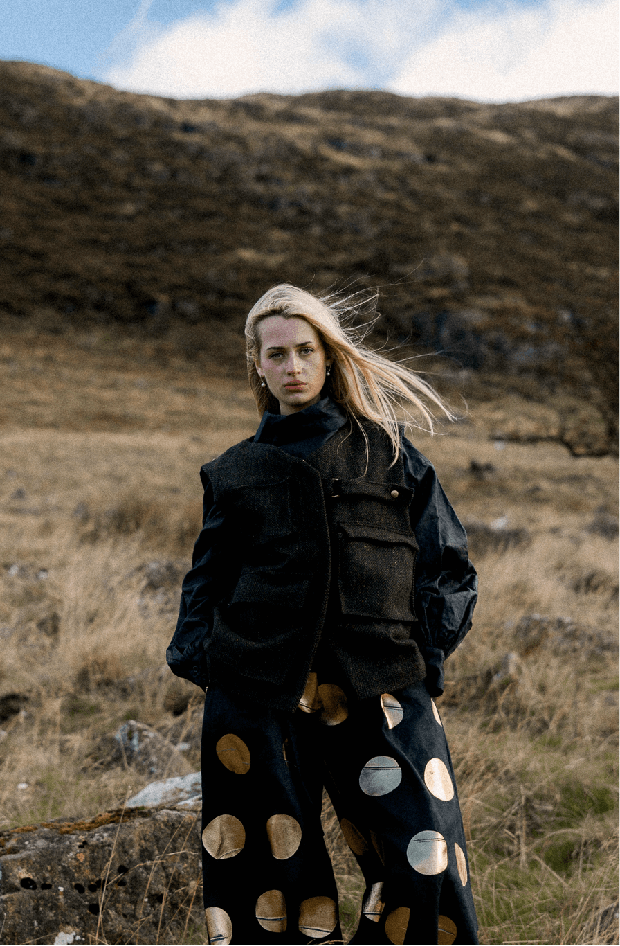 Model wearing a black blouse and black wide trousers with large gold circles, standing in a hillside landscape.