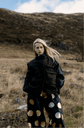 Model wearing a black blouse and black wide trousers with large gold circles, standing in a hillside landscape.
