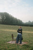 Woman in a dark jacket and wide trousers standing beside a chair in an open field under misty light.