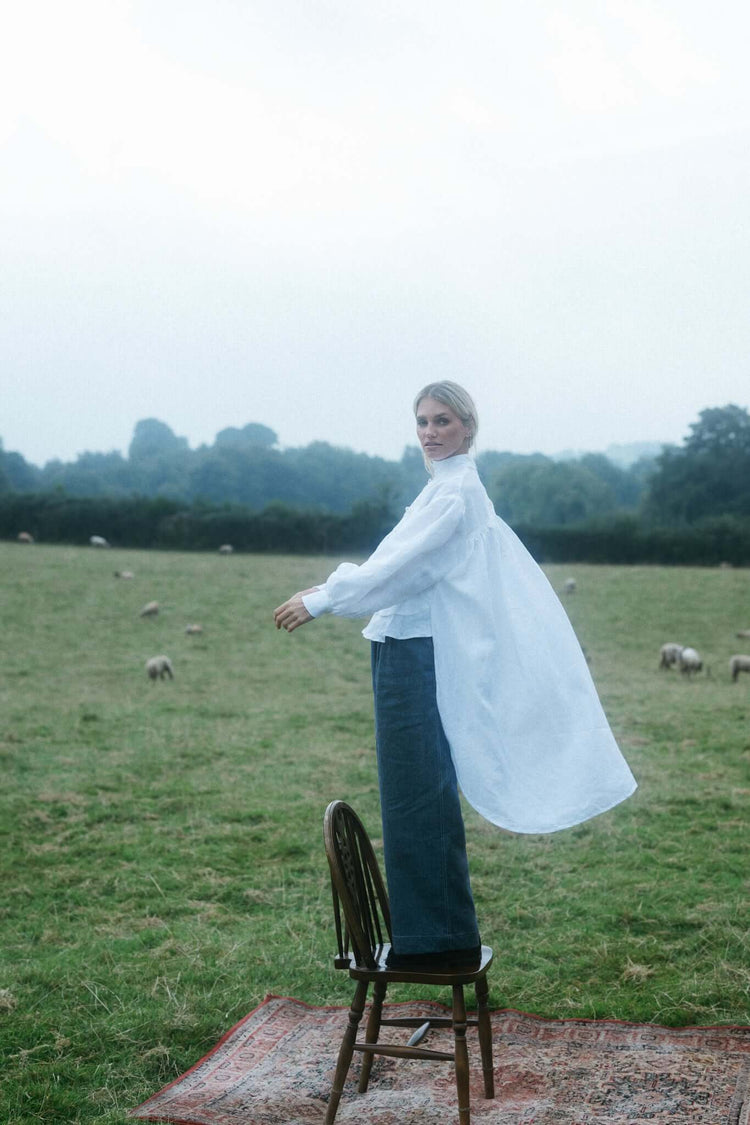Woman in a white cape-like top standing on a chair in an open field with sheep in the distance in soft natural light.