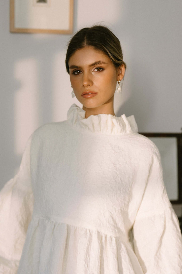 Woman in a white gathered blouse, photographed from the waist up in warm window light against a neutral wall.