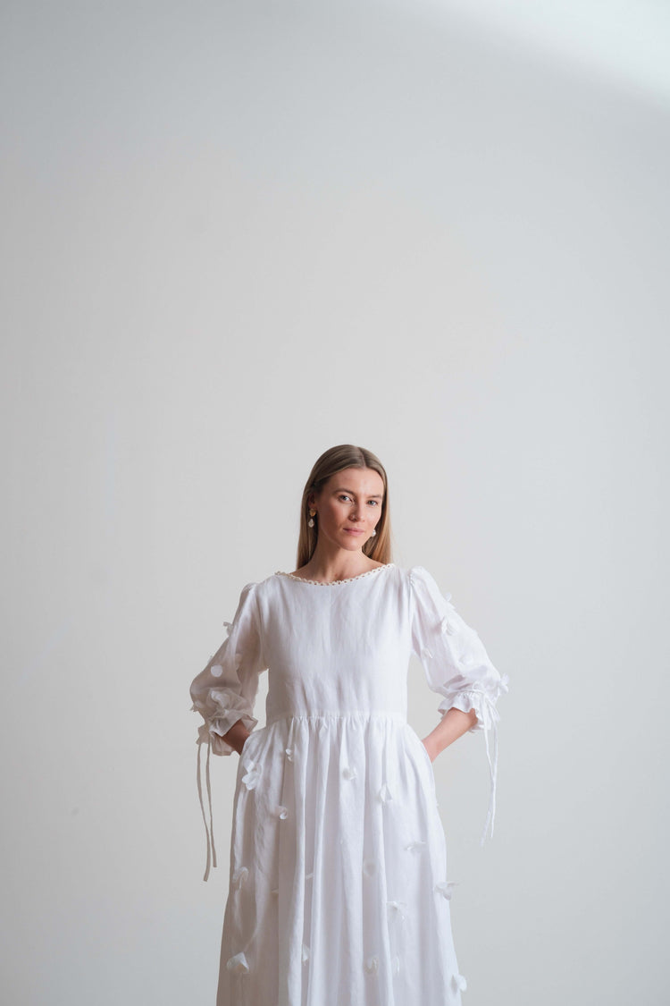White gathered dress with long sleeves and tie details, shown full length against a pale studio background.