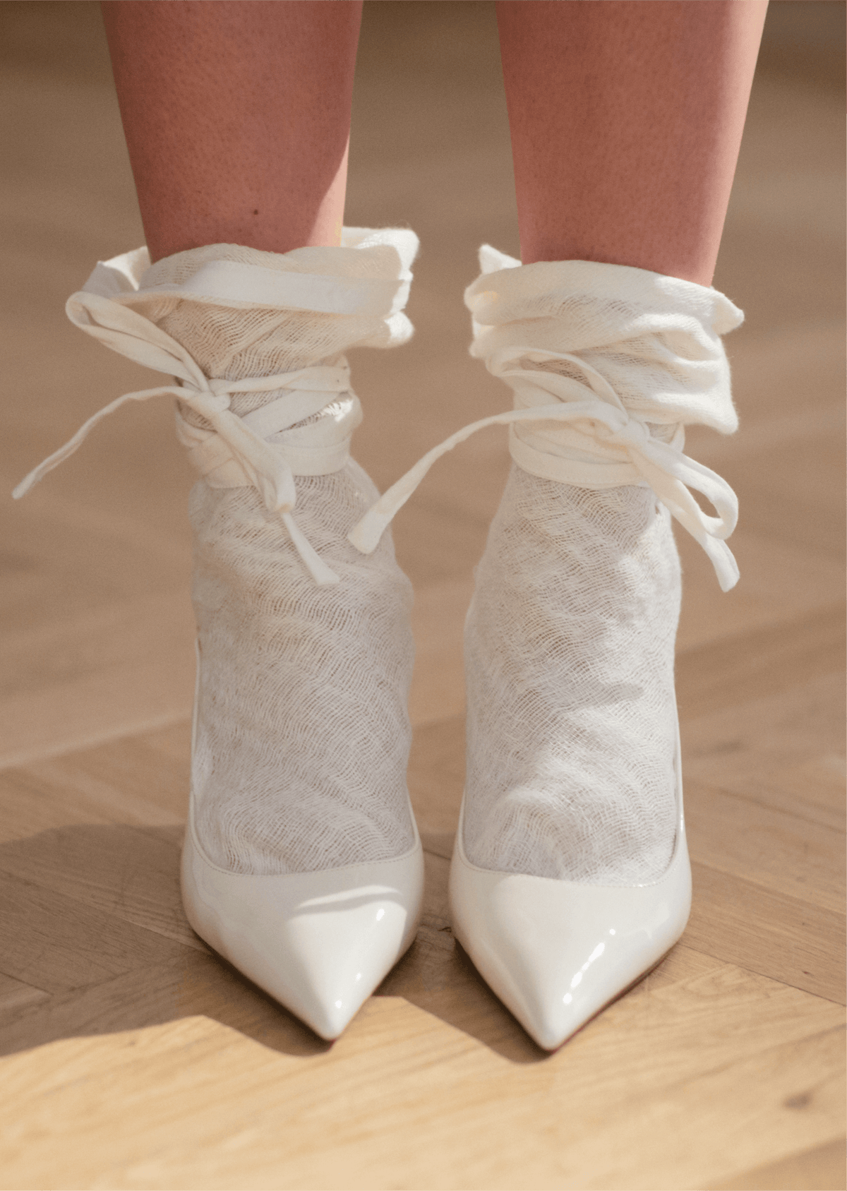 Close view of white pointed shoes with sheer lace ankle ties, photographed on a wooden floor in soft light.