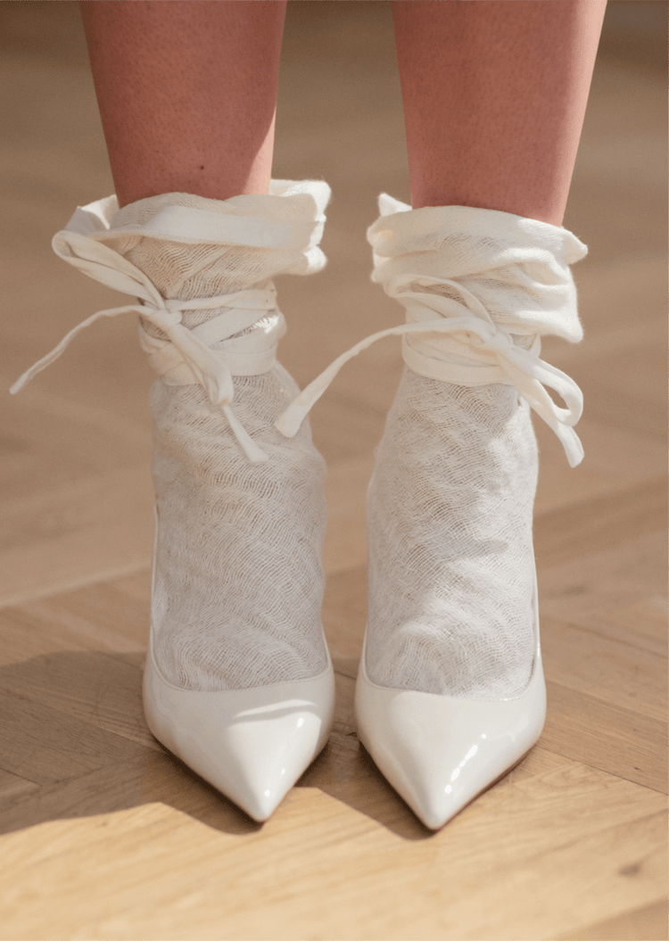 Close view of white pointed shoes with sheer lace ankle ties, photographed on a wooden floor in soft light.