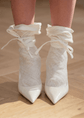 Close view of white pointed shoes with sheer lace ankle ties, photographed on a wooden floor in soft light.