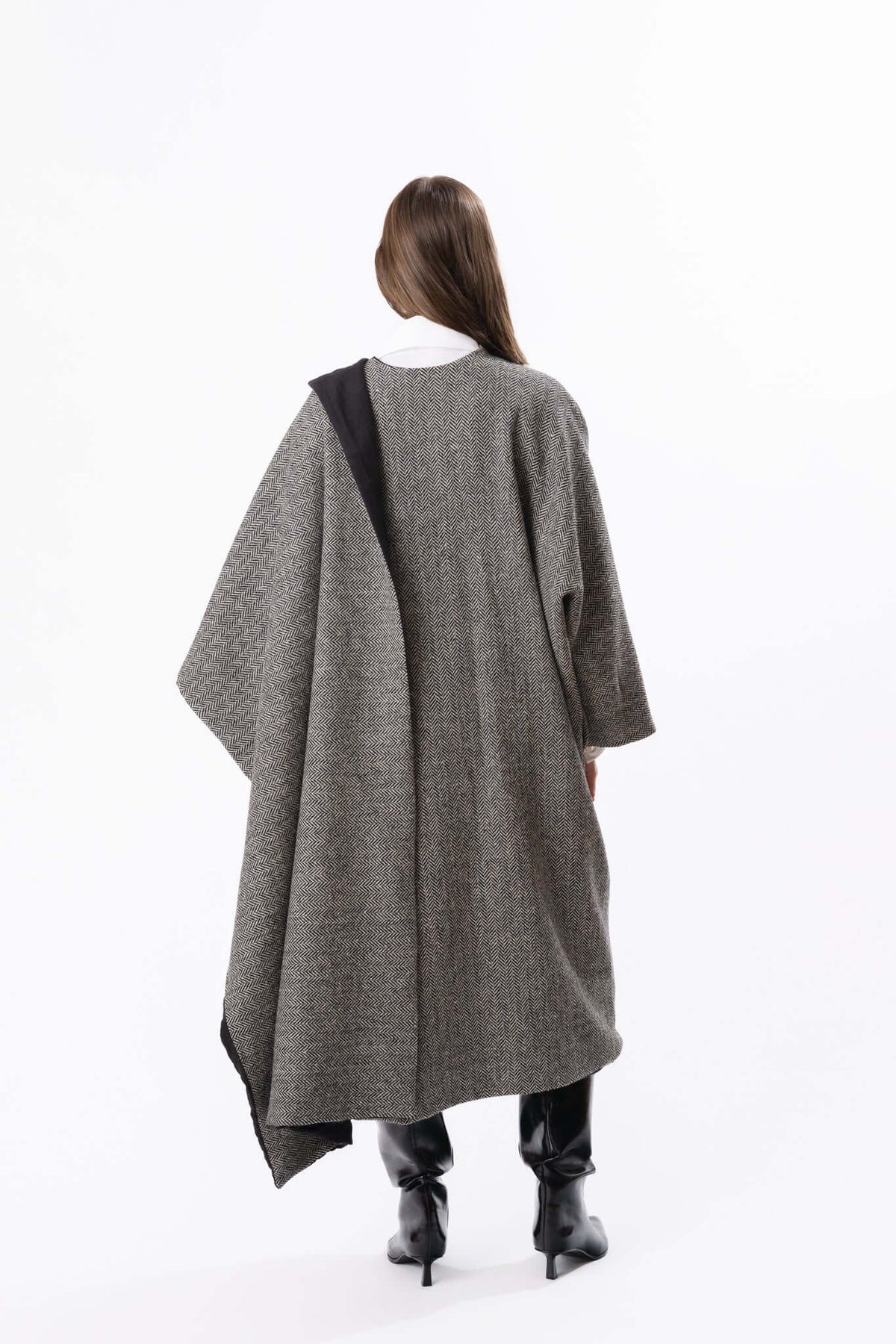 Back view of a grey cocoon-style cape dress worn with black boots against a plain studio backdrop in the studio.
