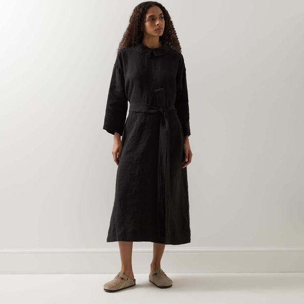 Woman in a dark belted dress standing against a neutral wall, photographed full length in soft indoor light.
