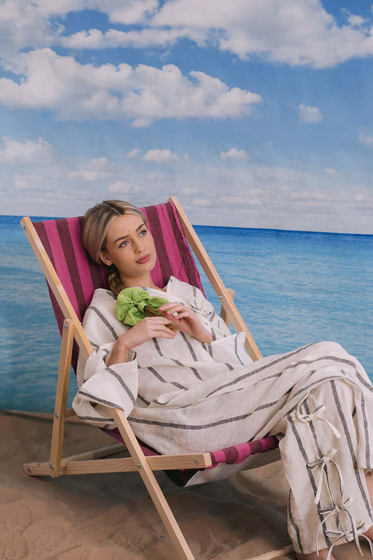 Woman reclining on a striped deckchair in a light outfit, photographed against a beach and sea backdrop.