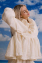Woman in a cream ruffled blouse, photographed against a beach backdrop with blue sky and bright sunlight.