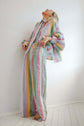 Woman in a pastel striped dress standing with head tilted back, photographed against a plain interior wall.