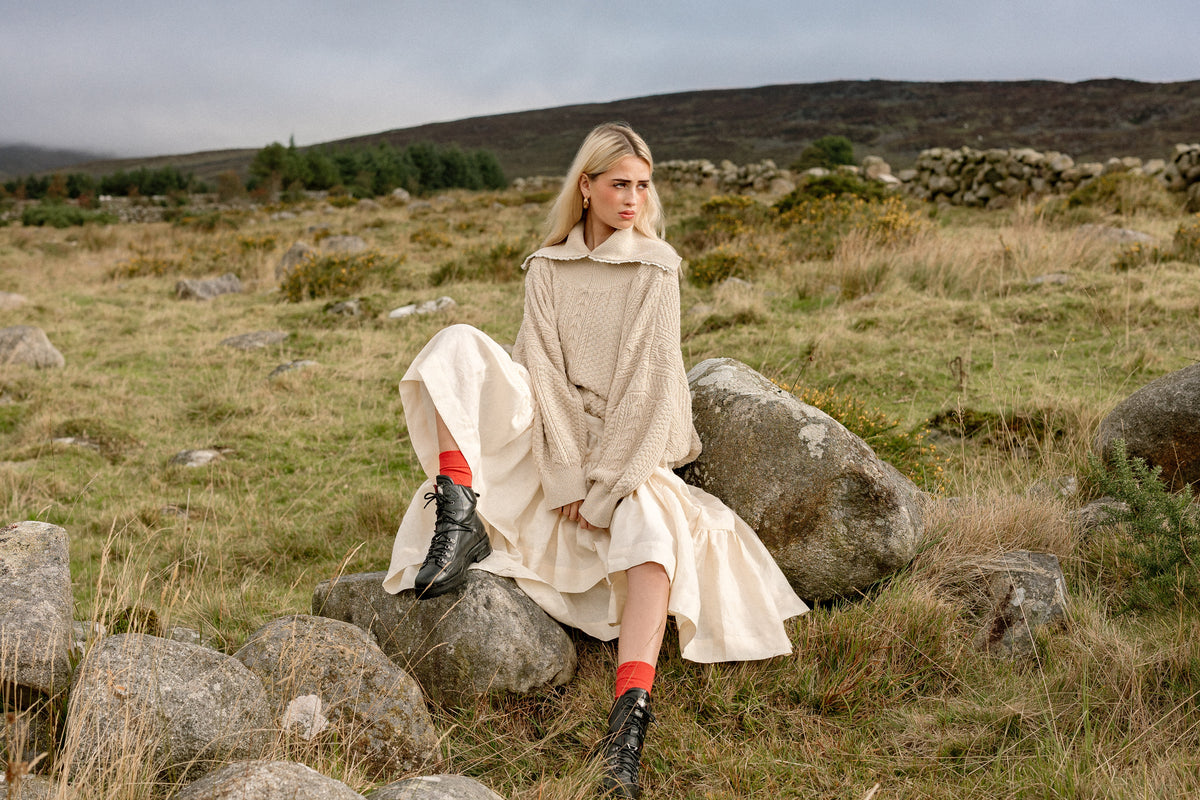 The Aran Jumper worn on the rocks of a hillside, with a cream linen skirt