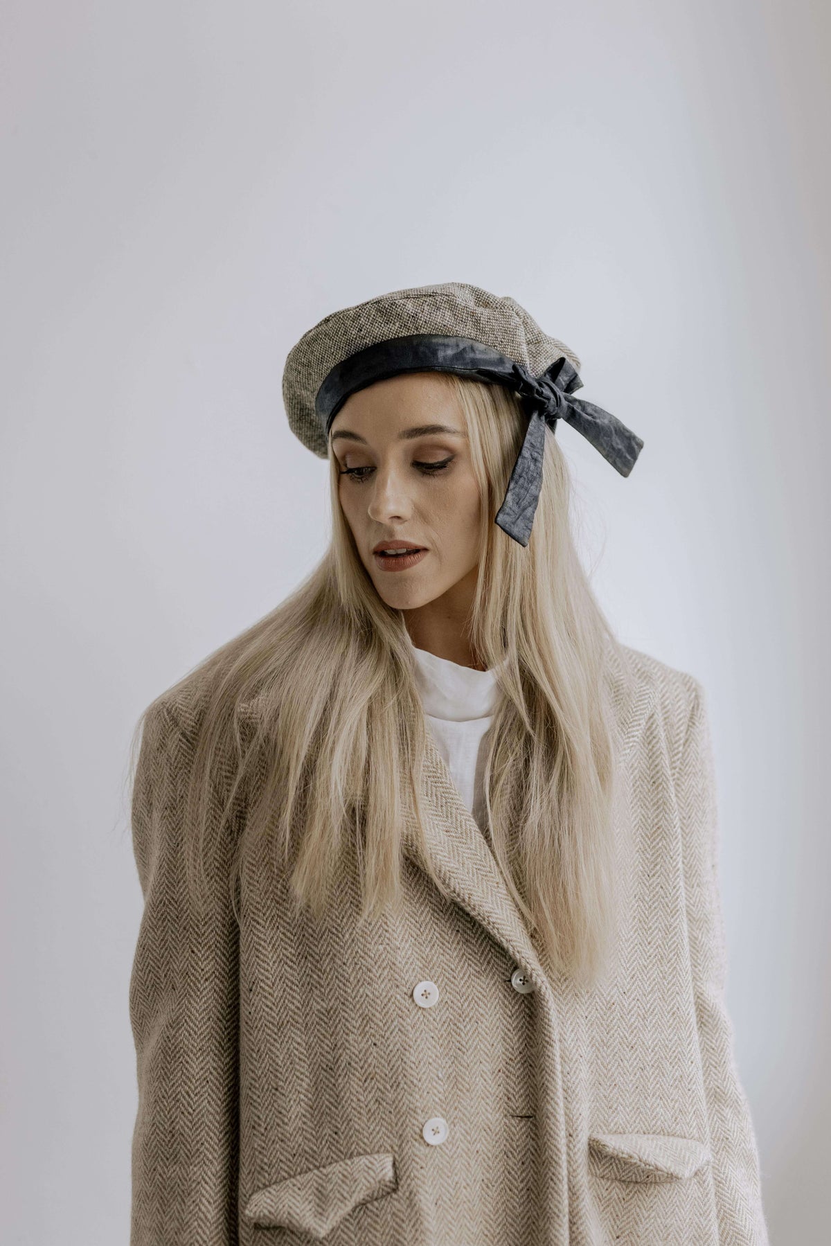 Portrait of a woman in a textured light coat and cap, photographed against a plain studio background.