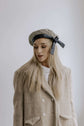 Portrait of a woman in a textured light coat and cap, photographed against a plain studio background.